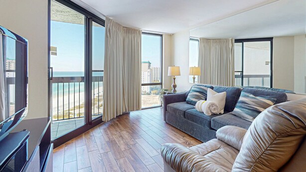A spacious living room with a large window, a sofa, a TV, and a balcony with a view of the city. - 16th floor condo with shared on-site pools, hot tub, restaurant, and bar (Destin)