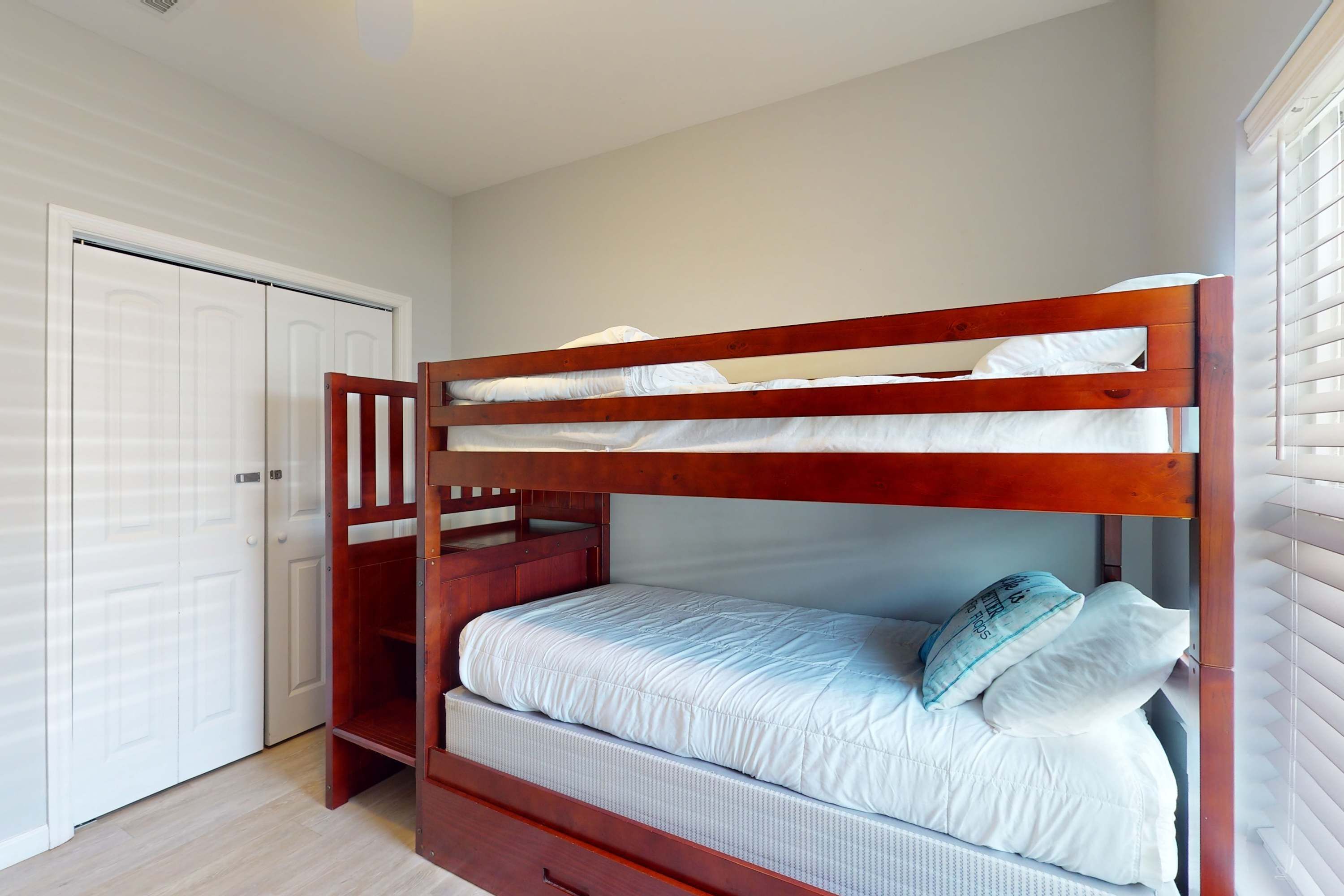 A bunk bed with a wooden frame and white bedding, a white door, and a window with blinds.