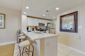 Une cuisine moderne avec des meubles blancs, un îlot central, des tabourets de bar, un grand miroir et des spots encastrés au plafond.