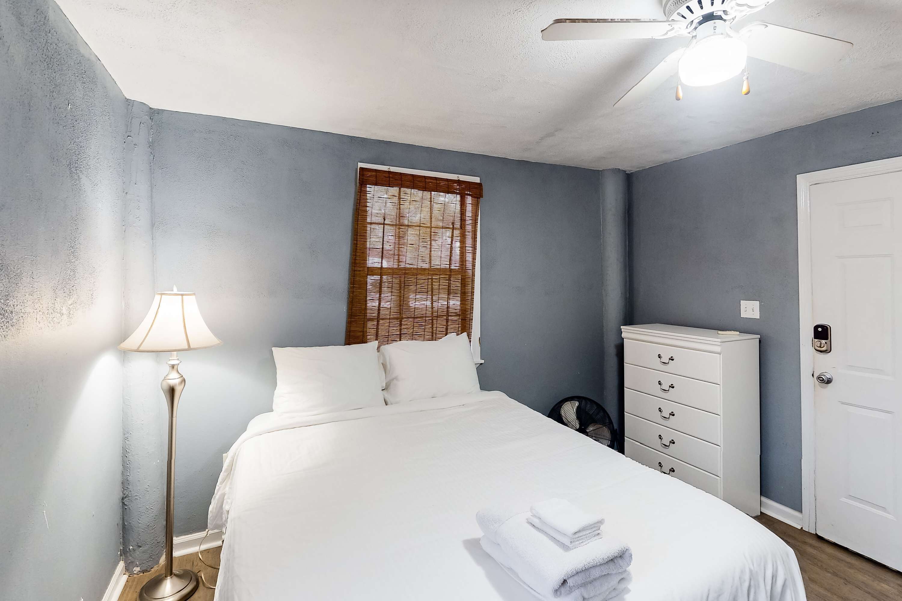 A bedroom with a bed, a bedside lamp, a dresser, a ceiling fan, and a window with blinds.