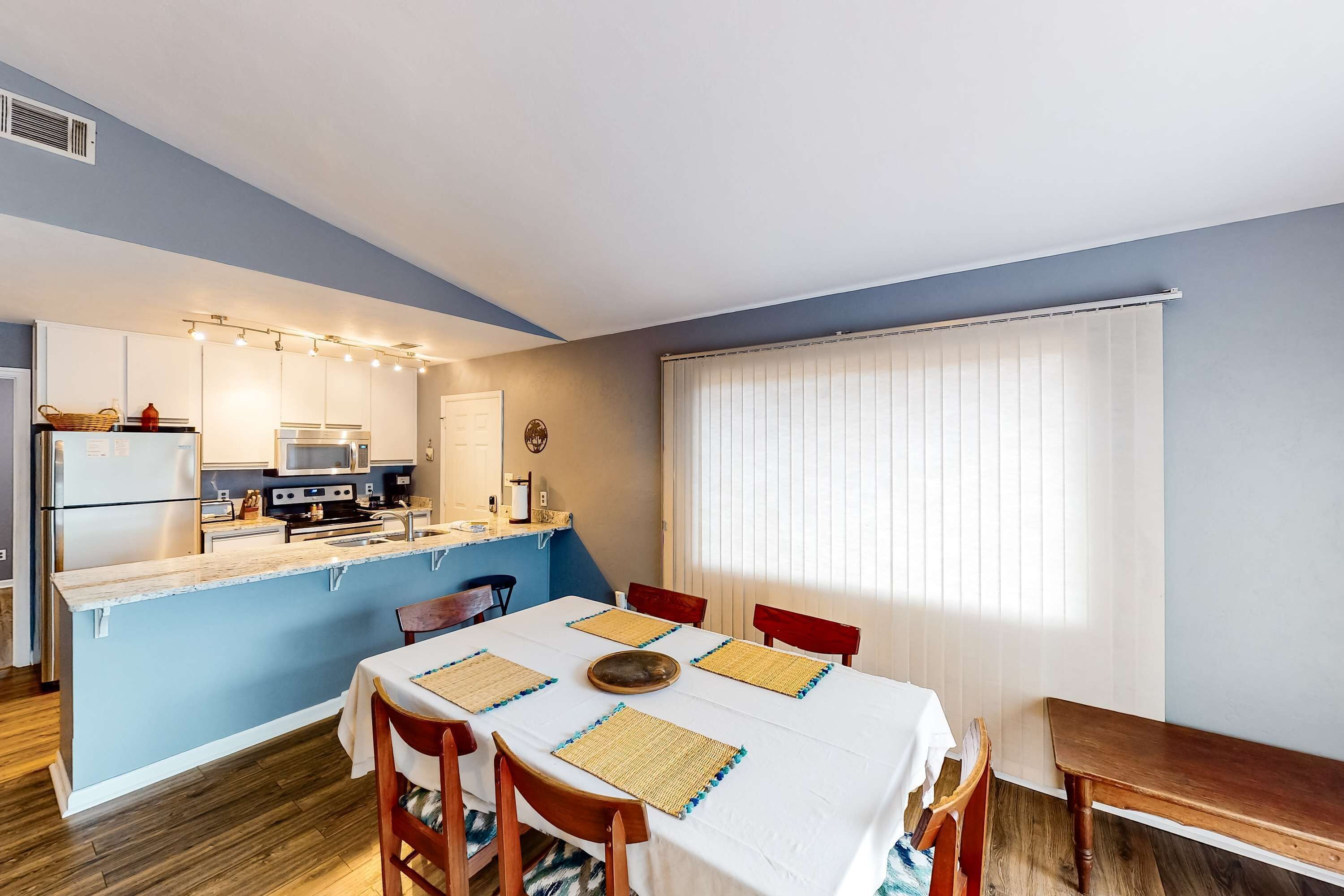 A compact kitchen with a dining area, featuring a central island, white cabinetry, and a window with blinds.