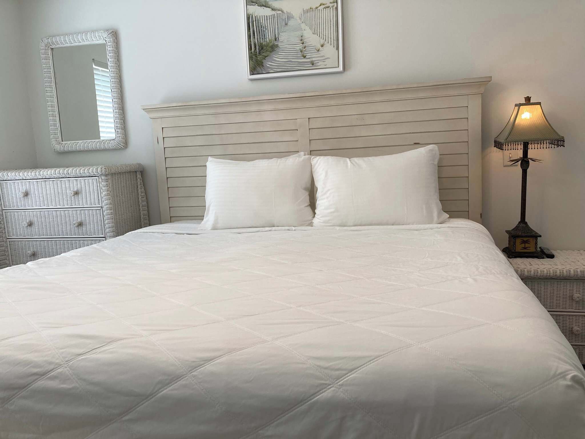 A neatly made bed with a quilted white duvet, a headboard with horizontal slats, a wall-mounted mirror, and a framed picture above the bed.