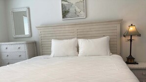Una cama bien hecha con un edredón blanco acolchado, un cabecero con listones horizontales, un espejo fijado a la pared y un cuadro enmarcado sobre la cama.