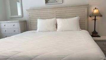 A neatly made bed with a quilted white duvet, a headboard with horizontal slats, a wall-mounted mirror, and a framed picture above the bed.