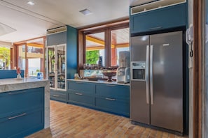 Une cuisine moderne avec des meubles bleus, un réfrigérateur en acier inoxydable et une grande fenêtre donnant sur un balcon.