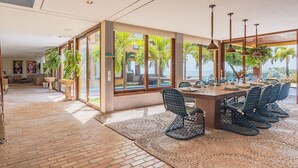 A spacious dining area with a long table, multiple chairs, and large windows offering a view of palm trees and the ocean.