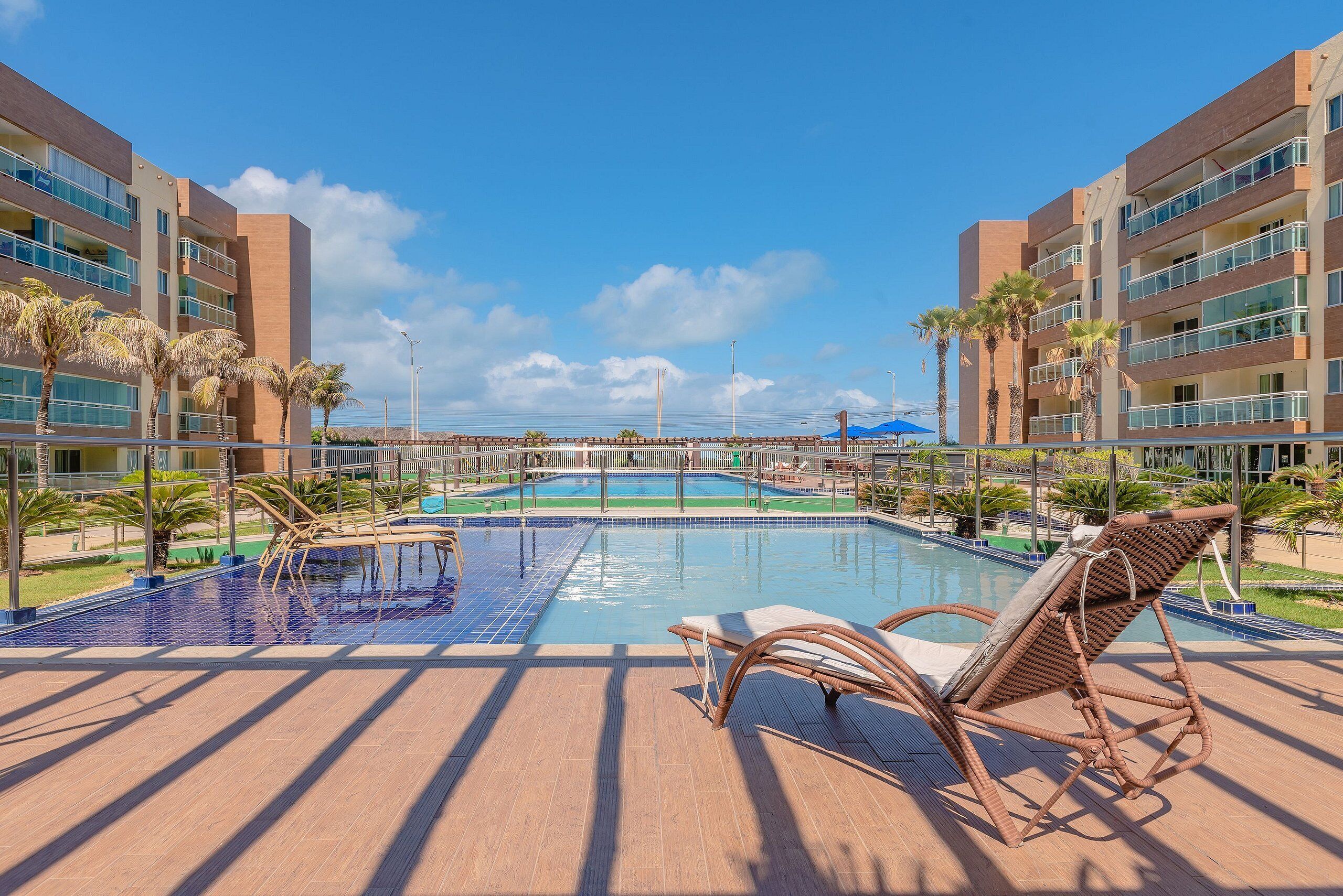 Uma área de piscina com espreguiçadeiras, céu claro e um edifício com varandas múltiplas.