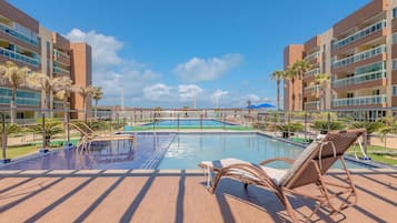 Uma área de piscina com espreguiçadeiras, céu claro e um edifício com varandas múltiplas.