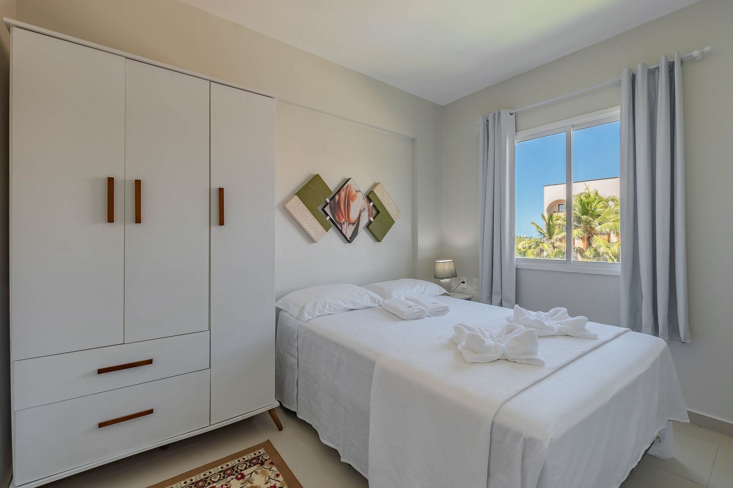 A bedroom with a bed, a wardrobe, a window with curtains, and a view of palm trees outside.