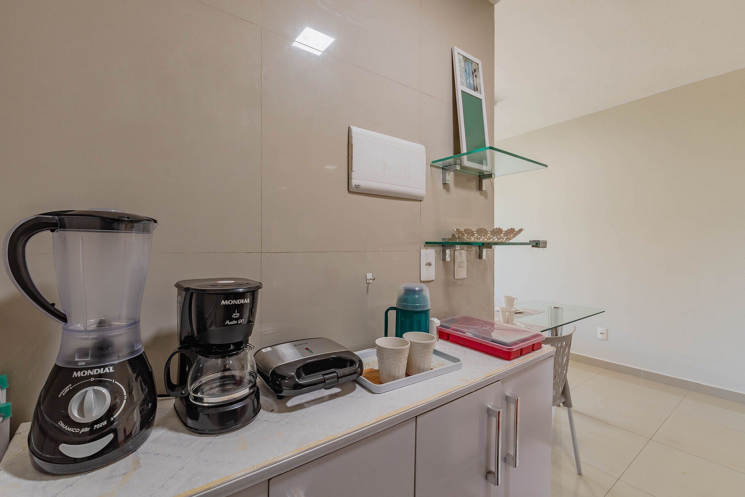 Una kitchenette con licuadora, cafetera y repisa de vidrio con una jarra de café y tazas.