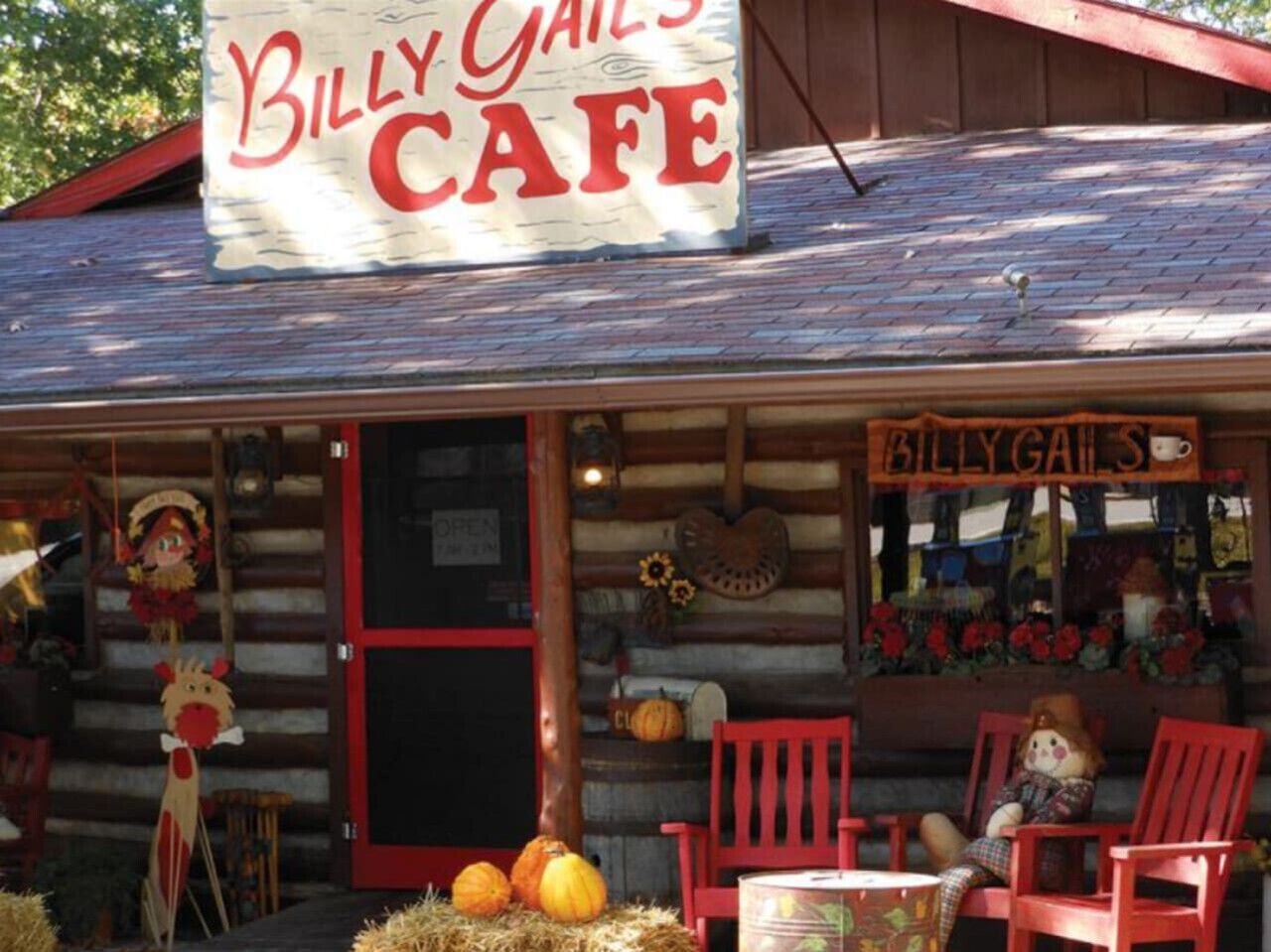 A rustic log cabin with a sign reading "Billy Gait's Cafe" and a porch with red chairs and pumpkins.