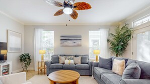 A living room with a grey sofa, a round wooden coffee table, a ceiling fan, and a large window with blinds. - Large, Open Home, Shared Pool, Bike or Walk to the Gulf (Destin)