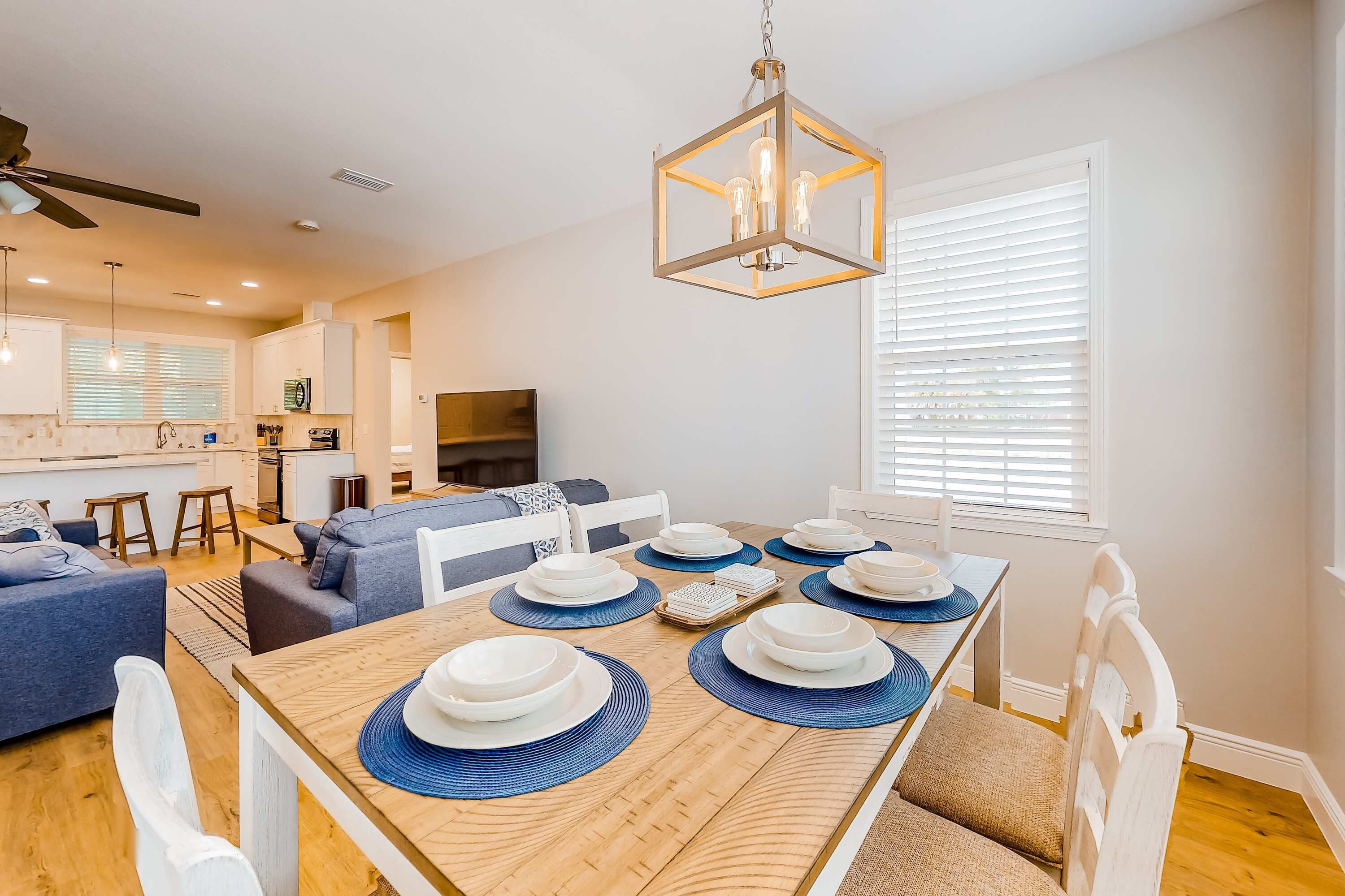 A modern dining area with a wooden table set for six, surrounded by white chairs. A ceiling fan and pendant light are visible above the table.