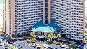 A high-rise hotel with multiple floors, a central courtyard, and a parking area with several cars. - Lovely Sixth-Floor Condo, with Shared Splash Pad, Multiple Pools, & Hot Tub (Destin)
