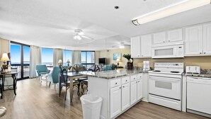 A modern kitchen with white cabinets, a microwave, and a dishwasher. There is a dining area with a table and chairs, and a view of the ocean through large windows. - Lovely Sixth-Floor Condo, with Shared Splash Pad, Multiple Pools, & Hot Tub (Destin)