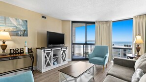 A hotel room with a large window, a flat-screen TV, a sofa, a chair, a small table, and a cabinet with a 'DREAM' sign. - Lovely Sixth-Floor Condo, with Shared Splash Pad, Multiple Pools, & Hot Tub (Destin)