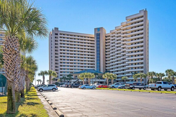 A multi-story hotel with a modern design, featuring a central atrium with a blue roof and a parking lot with several cars. - 14th Floor Airy Condo, On-site bar & restaurant, Pool w splash pad (Destin)
