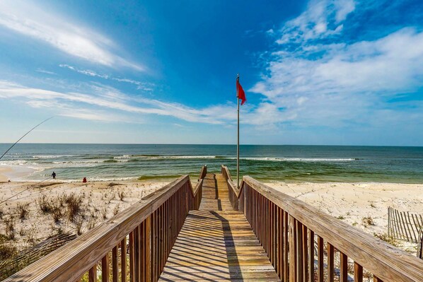 Une promenade en bois menant à une plage où flotte un drapeau rouge, avec un rivage sablonneux et des vagues déferlantes.
