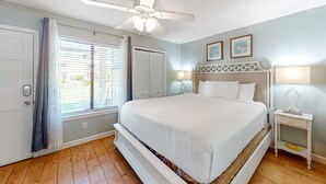 Un dormitorio con cama, ventilador de techo, ventana con persianas, mesita de noche y lámpara de pared.