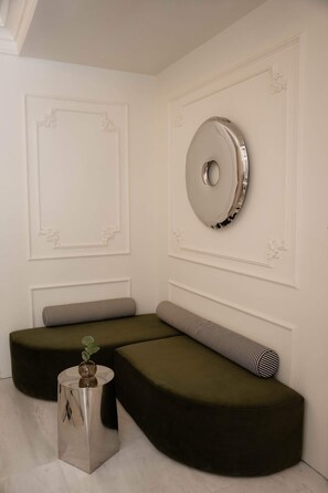 A room with a sectional sofa, a round mirror, and a small side table with a plant. - The Newel Kolonaki (Athens)