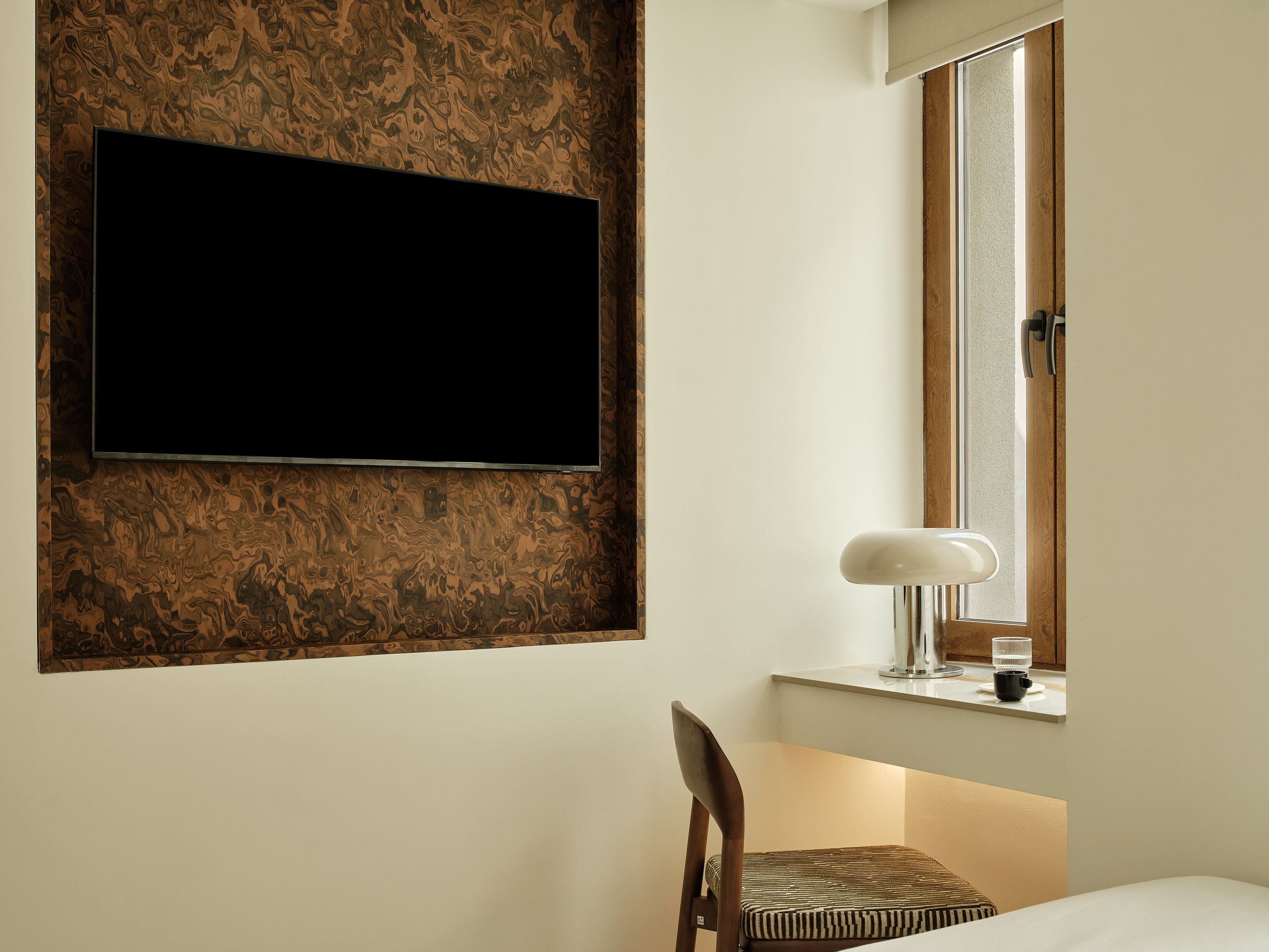 A room with a flat-screen TV mounted on a patterned wall, a window with a wooden frame, a bedside table with a lamp, and a chair with a striped cushion.
