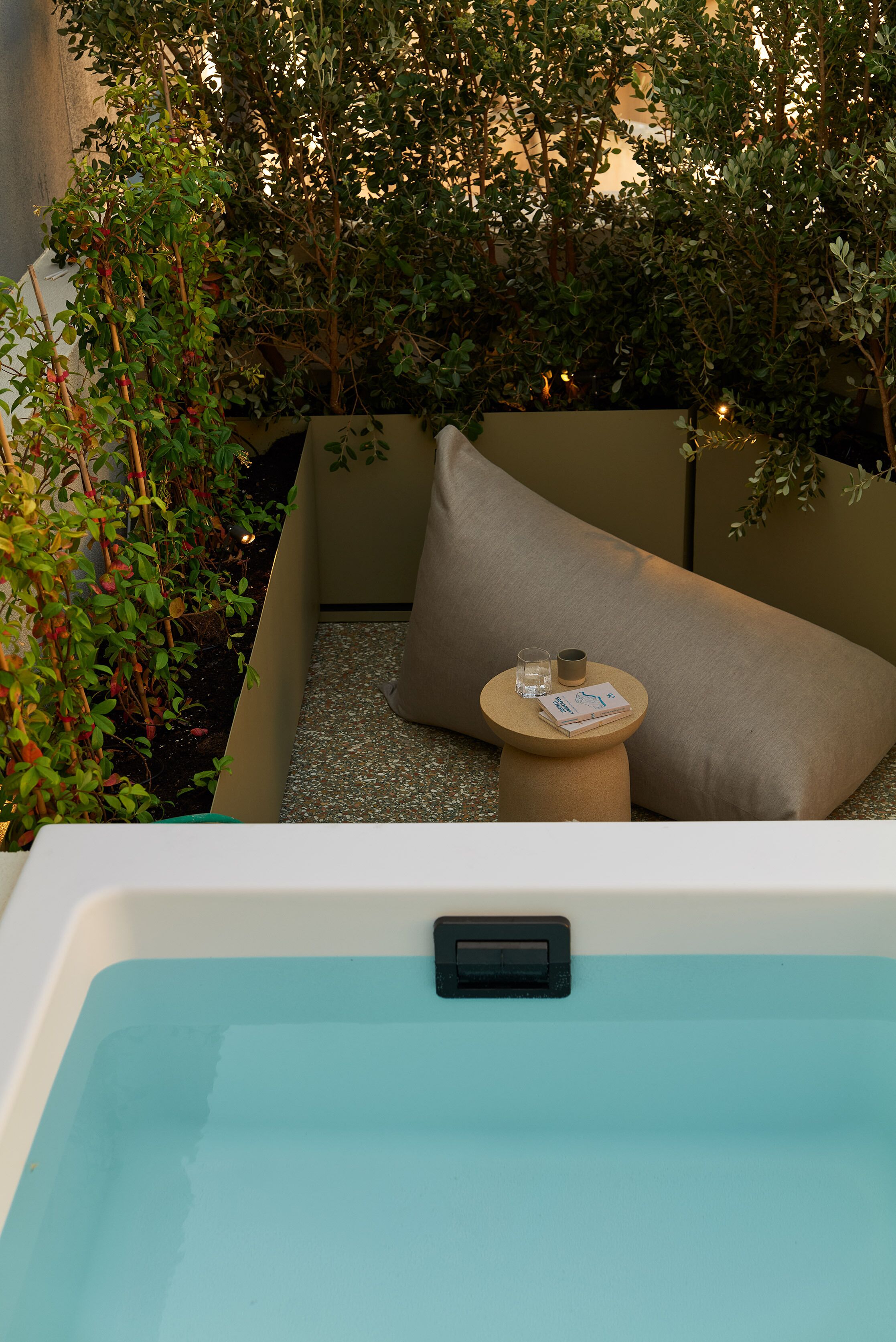 Un espacio de spa con un jacuzzi, una zona de estar acolchada y una pequeña mesa con una copa y un libro.
