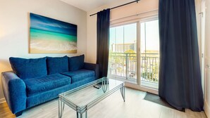 Een woonkamer met een blauwe bank, een glazen bijzettafeltje, een schilderij van een strand en een balkon met uitzicht op gebouwen.