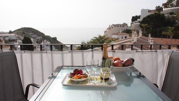 Et bord med champagneflaske, glas og et frugtfat på en balkon med udsigt over en kystby.