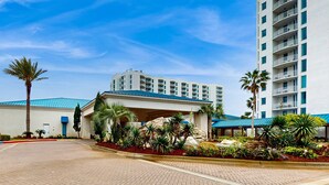 Ein Hotel mit blauem Dach, überdachter Eingangspartie und einem angelegten Garten mit Palmen und anderen Pflanzen.