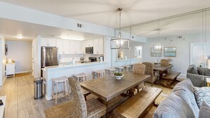 Uma cozinha moderna com eletrodomésticos de aço inoxidável, uma sala de jantar com mesa de madeira e cadeiras de vime, e uma sala de estar com sofá e mesa de centro.