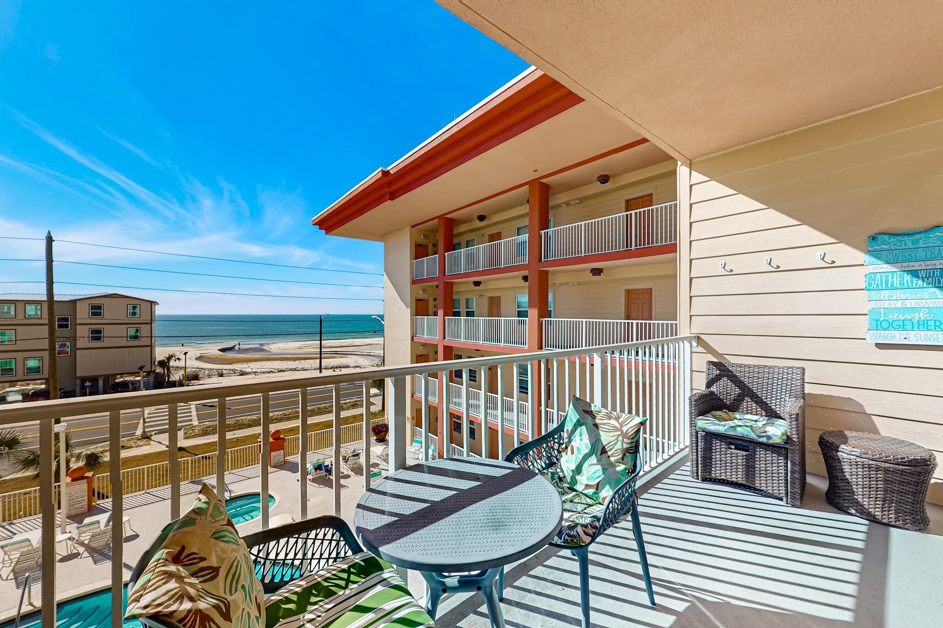 A balcony with a table and chairs, overlooking a beach and a building with a sign that reads "GATHER FAMILY TOGETHER TO WATCH THE SUNSET."