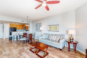 A living room with a sofa, coffee table, and chairs. There is a kitchen area with cabinets and a dining table. A ceiling fan is visible. - Gulf-view condo with outdoor pool, hot tub, gym & tennis - walk to the beach (Destin)