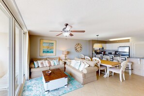 A living room with a sofa, coffee table, and dining area with chairs. There is a ceiling fan, a clock, and a kitchen area with appliances. - Lake-view 5th-floor resort condo with outdoor pool, gym, balcony, & washer & dryer (Destin)
