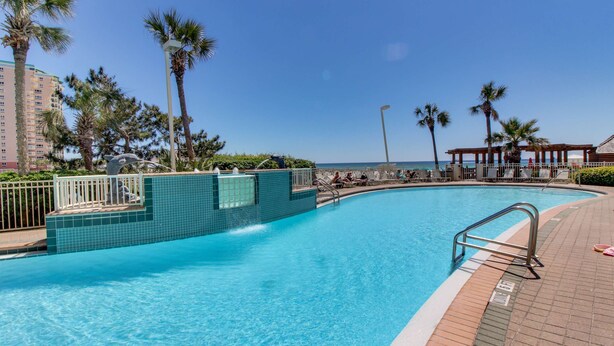 Une piscine dont le bord est revêtu de carrelage, entourée de palmiers et surplombée d’un ciel bleu azur.