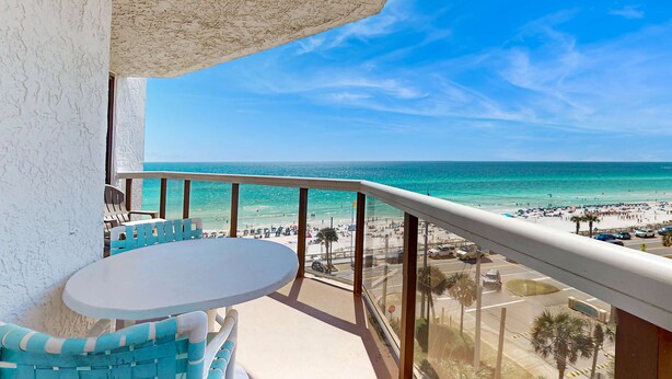 Ein Balkon mit rundem Tisch und blauen Stühlen, der Blick auf einen Strand mit Menschen und Autos geht.