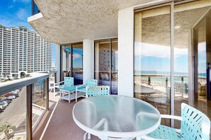 En balkon med et glasbord og stole, der kigger ud over en strand og en parkeringsplads.