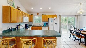 A kitchen with wooden cabinets, a granite countertop, a dining area with chairs, and a view through a glass door. - Dog-friendly home near the beach with spacious living area, porch & sundeck (Port St. Joe)