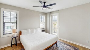 Une chambre à coucher avec un lit, un ventilateur de plafond, une fenêtre avec des stores, une petite table avec une lampe et un tapis à motifs.