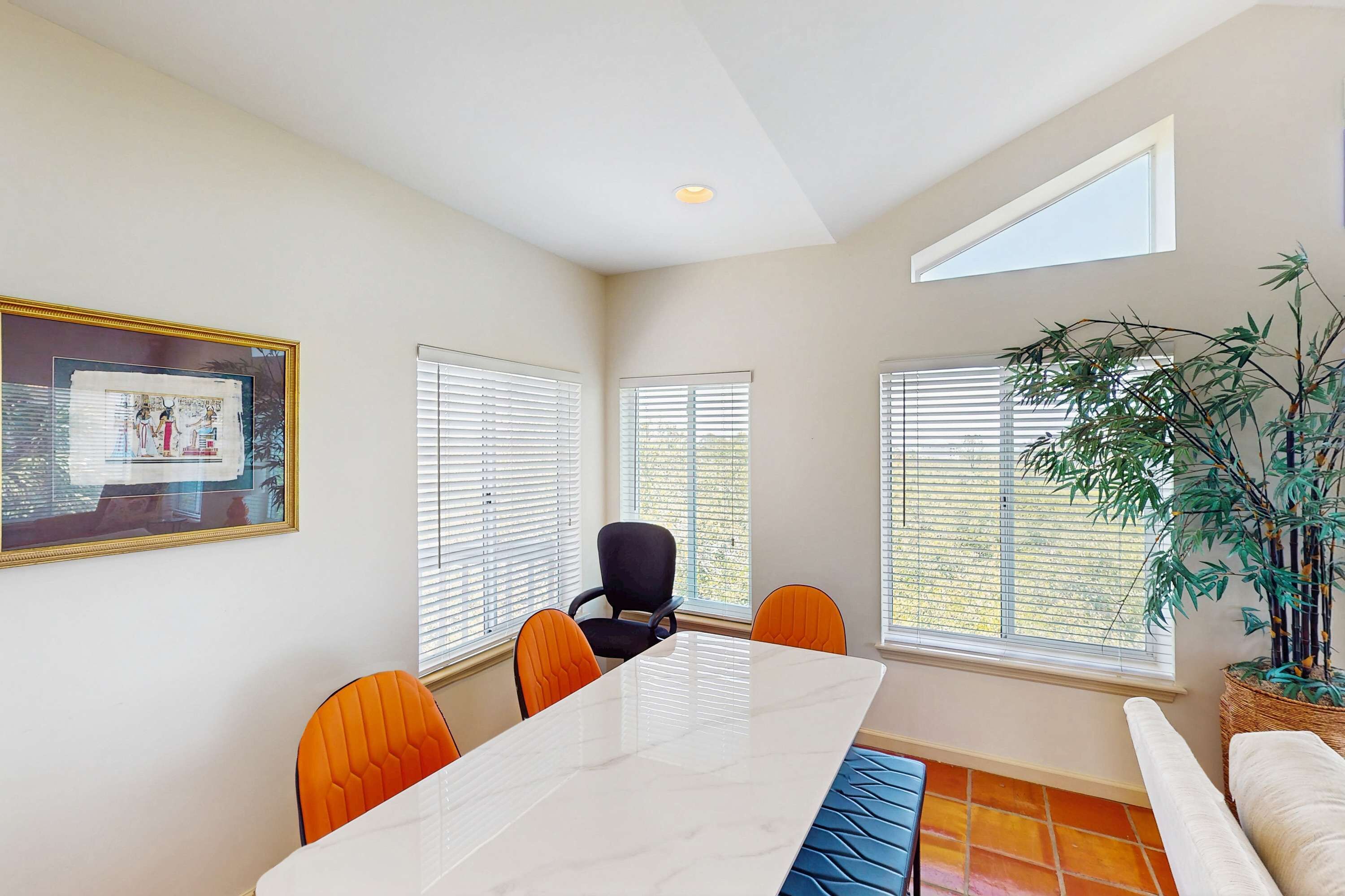 A dining area with a marble table, orange chairs, a black chair, and a large potted plant.