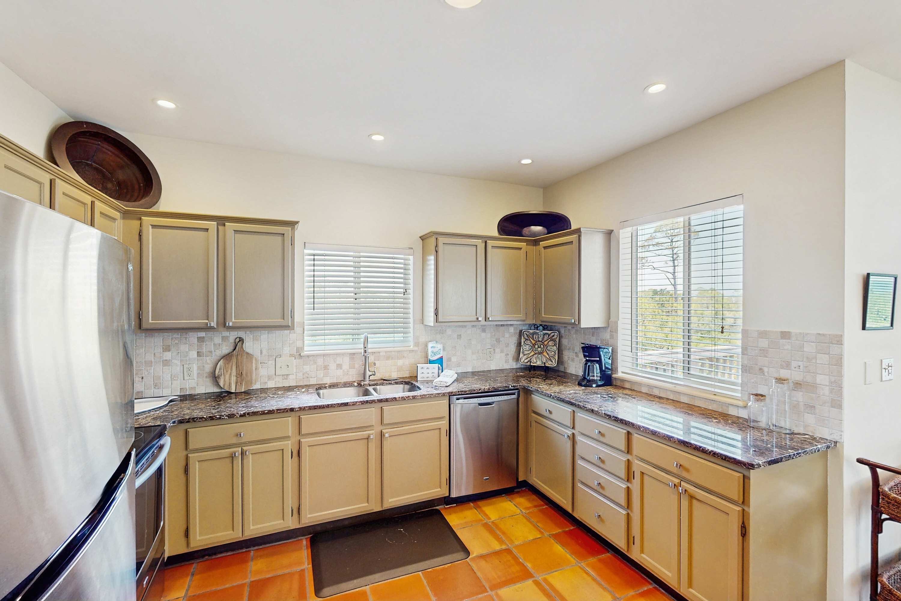 A kitchen with beige cabinets, a stainless steel refrigerator, a dishwasher, and a window with blinds.