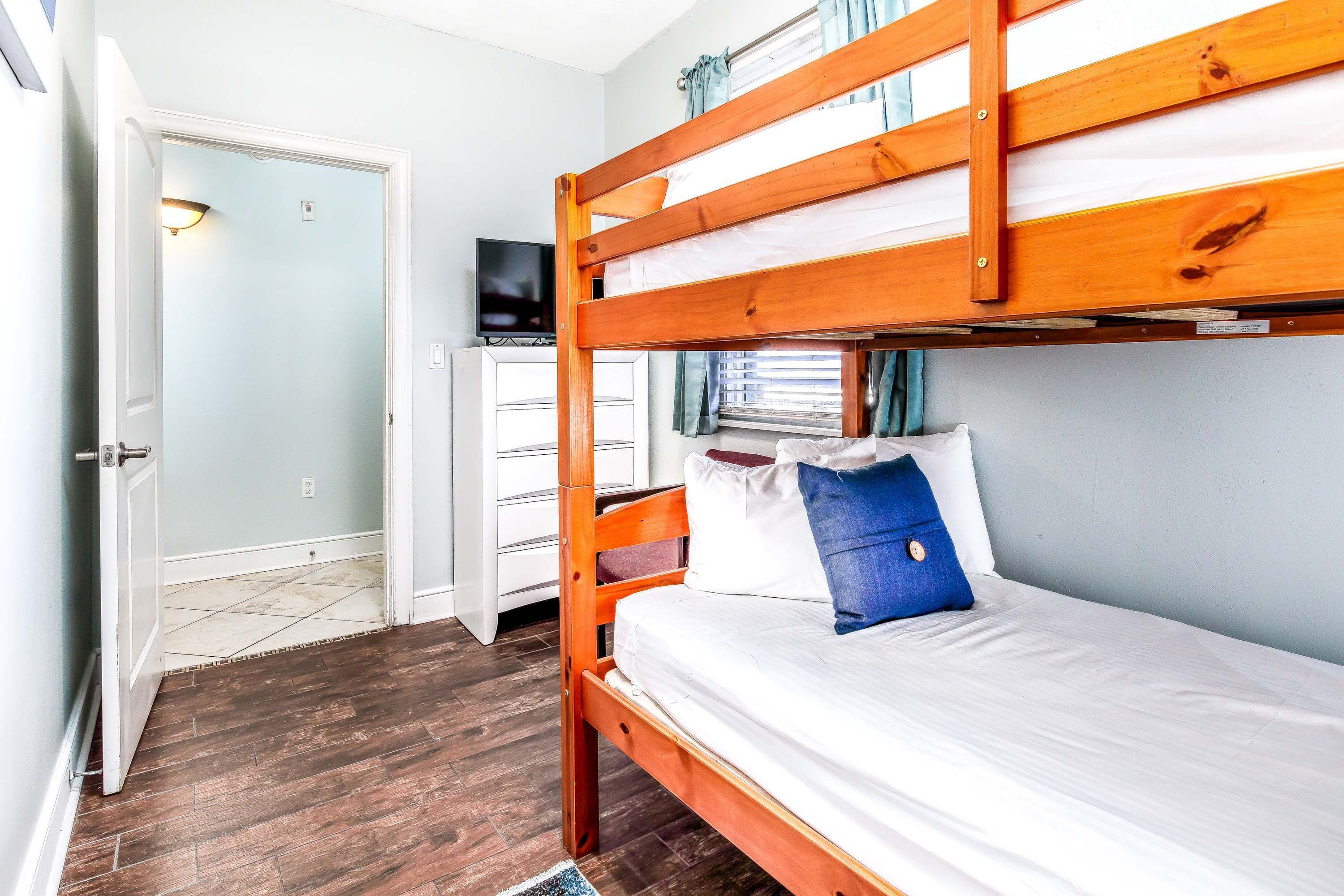 Een slaapkamer met een stapelbed en een televisie op een dressoir, een deur die naar een andere kamer leidt, en een raam met gordijnen.