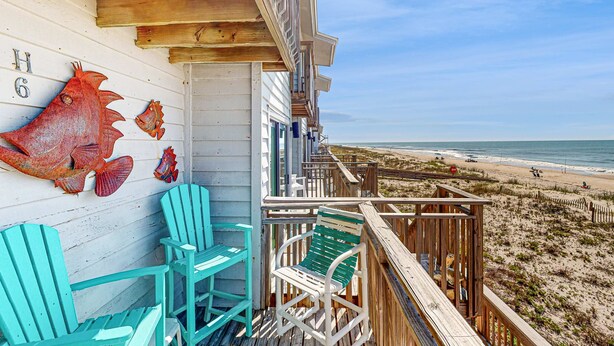 Une terrasse en bois avec des chaises turquoise, une rambarde en bois et une vue sur la plage.