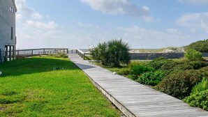 Une promenade en bois mène à une plage, avec une zone herbeuse et quelques buissons d’un côté.