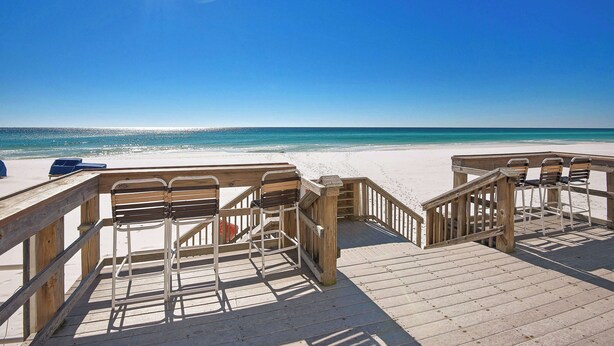 Une terrasse en bois, dotée de chaises en métal et d’une rambarde en bois, qui offre une vue sur la plage.