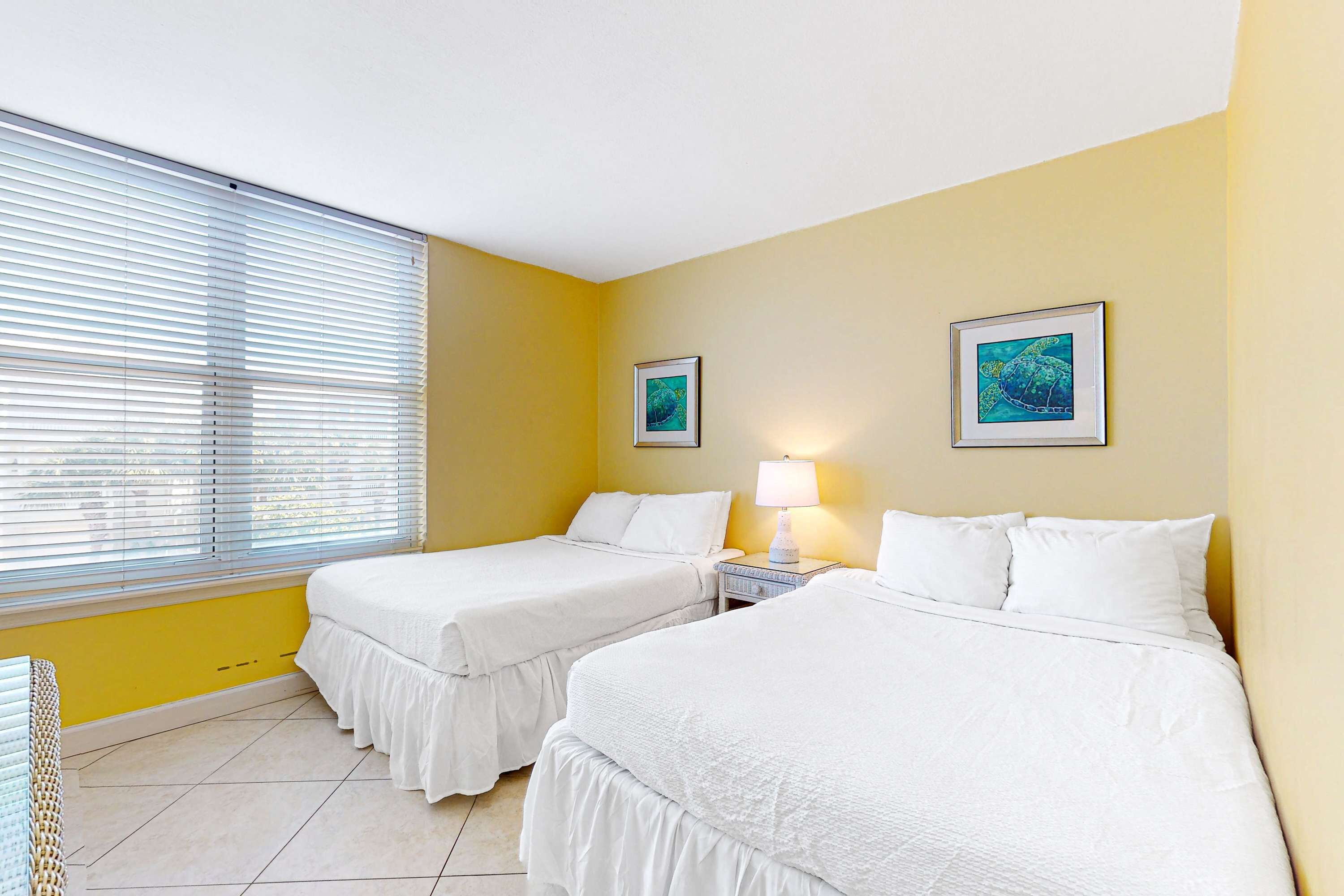 A hotel room with two beds, a window with blinds, and two framed pictures on the wall.
