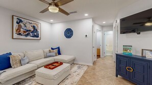 Uma sala de estar com sofá seccional, mesa de centro, ventilador de teto, televisão na parede e um armário com objetos decorativos.