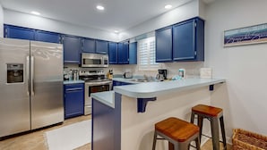 Uma cozinha moderna com armários azuis, eletrodomésticos de aço inoxidável, uma ilha central com banquetas e uma placa de boas-vindas.