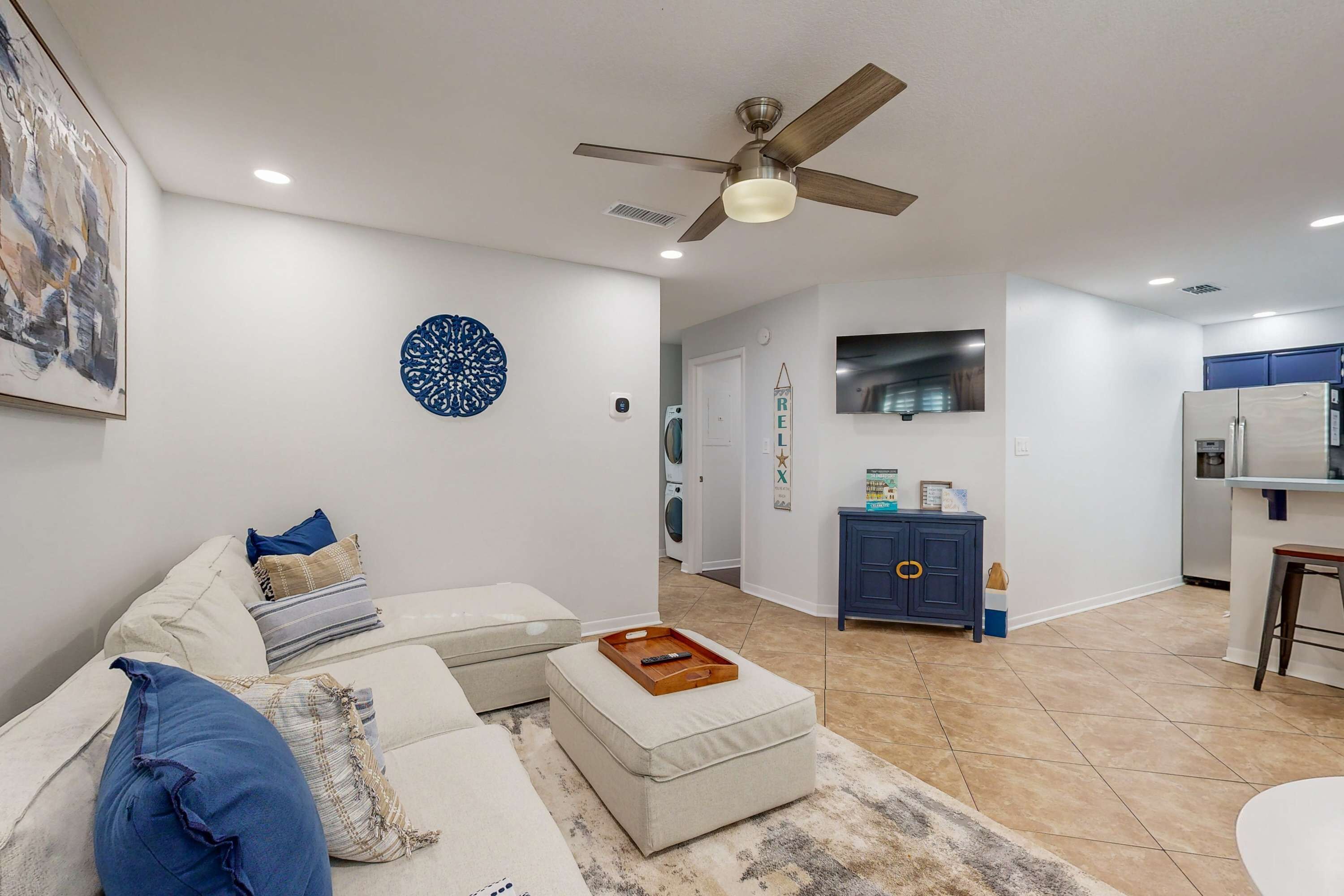 Un salon moderne avec un canapé, un pouf et un ventilateur de plafond. On aperçoit à l’arrière-plan un coin cuisine avec un réfrigérateur et des placards.