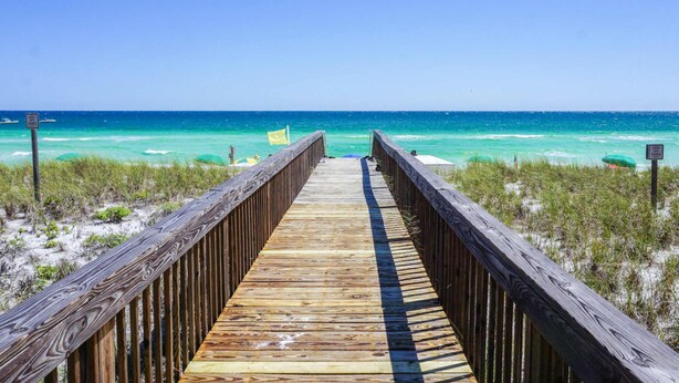 Een houten looppad dat naar een strand leidt met helder turquoise water en groene begroeiing.