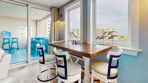 Een eetgedeelte met een houten tafel en vier stoelen, grote ramen met uitzicht op de stad, en een balkon met blauwe stoelen.